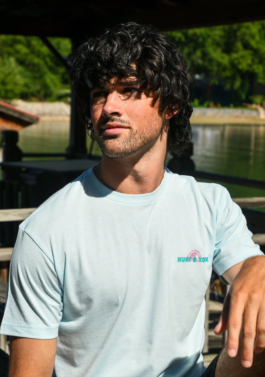 Man wearing a light blue t-shirt with a logo, sitting outdoors near water.
