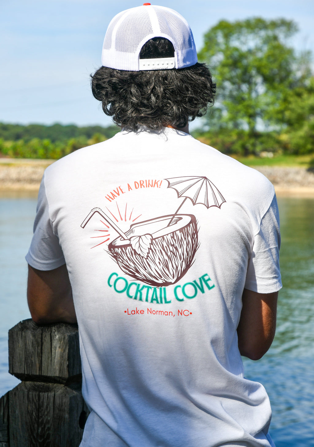 Person wearing a white t-shirt with 'Cocktail Cove' design by a body of water.